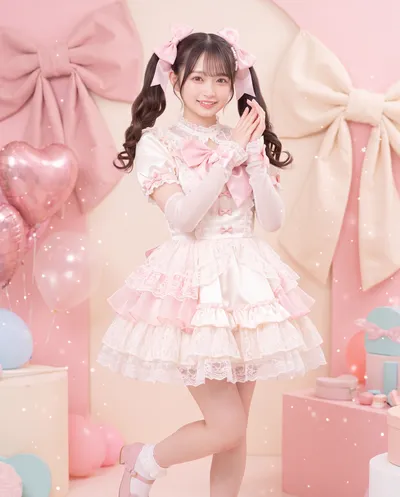Rino in a frilled cream and pink dress with long gloves and hair bows, hands clasped among giant bows and heart balloons